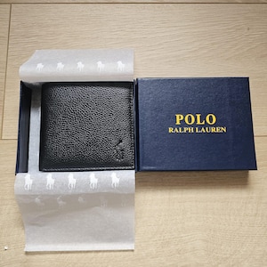 May include: A black pebbled leather bifold wallet featuring an embossed pony logo, presented in a navy blue box with the yellow text POLO RALPH LAUREN. The wallet rests on white tissue paper patterned with repeating white pony logos.