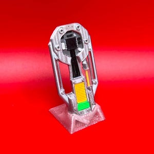 May include: A silver-colored, vertical, mechanical-style prop standing on a triangular base. The structure features a central black electronic component, a yellow and green textured panel, and a thin orange vertical bar, all encased in a frame.