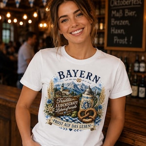 Puede incluir: Una camiseta blanca con un estampado gráfico colorido de los Alpes bávaros, una jarra de cerveza, un pretzel y espigas de trigo. El texto dice BAYERN, MIA SAN MIA, Tradition Leidenschaft Lebensfreude y PROST AUF DAS LEBEN! con detalles azules.