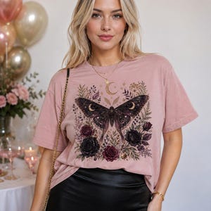 Op de afbeelding: Een oudroze T-shirt met korte mouwen en een centrale grafische print van een grote donkere nachtvlinder met maansikkelmarkeringen op de vleugels, omringd door gedetailleerde illustraties van donkere rozen, gebladerte en een gouden maansikkel.