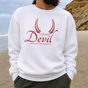 May include: A white crewneck sweatshirt featuring a red graphic of two devil horns above the text "I AM THE Devil AND DON'T YOU FORGET IT" printed in a red serif font across the centre of the chest, with long sleeves and ribbed cuffs.