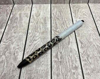 May include: A black and gold glitter pen with a leopard print design. The pen is on a white wood background.