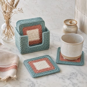 May include: A set of square crochet coasters featuring a teal border, a dusty rose middle band, and a cream centre. The coasters are stored in a matching teal crochet holder, with one coaster placed flat and another supporting a ceramic mug.