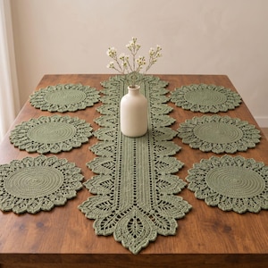 May include: A sage green crochet table runner with a decorative leaf-patterned border and six matching circular crochet placemats. A smooth, off-white ceramic vase containing a small bouquet of white flowers sits in the centre of the runner.