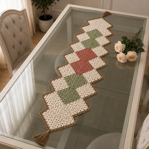 May include: A rectangular crochet table runner featuring a geometric pattern of cream, sage green, and dusty rose squares. The runner is bordered in brown yarn and finished with a brown tassel at each end, creating a long, decorative textile piece.