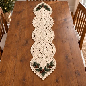 May include: A long, cream-colored crochet table runner featuring a repeating leaf-like pattern with scalloped edges. The ends are decorated with dark green embroidered pine needles and small brown pinecone accents in a symmetrical arrangement.