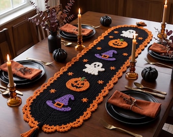 May include: A black crocheted table runner featuring orange pumpkin, purple witch hat, and white ghost motifs. The runner has an orange scalloped border and black and orange tassels at each end, displayed on a wooden dining table with place settings.