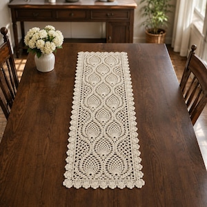 May include: A rectangular cream-colored crochet table runner featuring a repeating pineapple-inspired motif with an intricate openwork pattern and a scalloped edge, centered on a dark brown wooden table surface.