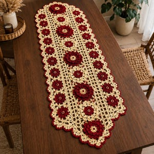May include: A long, rectangular crochet table runner featuring a cream-colored lattice pattern accented with deep red, three-dimensional floral motifs arranged in a central row and along the scalloped edges of the textile piece.