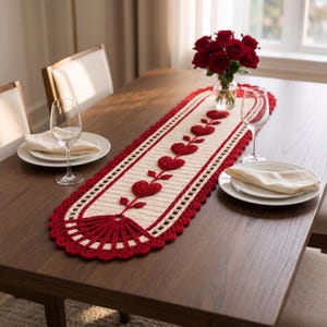 May include: An elongated, cream-colored crochet table runner featuring a central row of red heart motifs connected by stems. The runner has a scalloped red border and a fan-shaped crochet design at each end, creating a decorative textile piece.