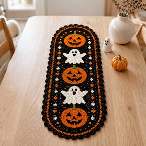 Puede incluir: Un camino de mesa ovalado de ganchillo con fondo negro, que alterna calabazas de Halloween naranjas y fantasmas blancos. El camino tiene un borde negro festoneado y pequeños detalles florales en blanco y naranja.