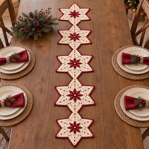 Puede incluir: Un camino de mesa de ganchillo en color crema y rojo oscuro con cinco motivos en forma de estrella conectados con centros florales. El camino se muestra sobre una mesa de madera con cuatro servicios, cada uno con plato y servilleta roja.