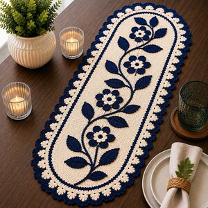 May include: An oval-shaped cream-colored crochet table runner featuring a dark navy blue floral vine pattern with five flowers and several leaves. The runner has a scalloped border in cream and navy blue, creating a decorative textile accent.