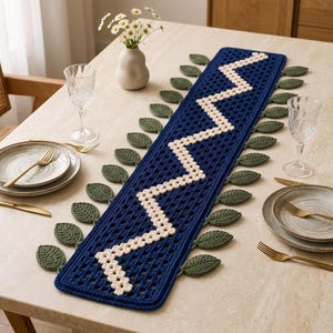 Puede incluir: Un camino de mesa de ganchillo rectangular con un patrón de rejilla azul oscuro, un diseño central en zigzag de color crema y hojas de ganchillo de color verde oscuro decorativas unidas a lo largo de los bordes largos.