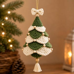 Op de afbeelding: Een kegelvormige kerstboomversiering gemaakt van gehaakt garen in afwisselende lagen van bosgroen en crème. Het ontwerp is voorzien van kleine houten kralen tussen de lagen, een gehaakt strikje aan de bovenkant en een crèmekleurig kwastje aan de onderkant.