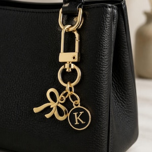 May include: A black textured handbag featuring a gold-toned metal keychain attachment. The keychain consists of a gold bow charm and a circular black pendant with the letter K in gold, suspended from a gold lobster clasp and ring hardware.