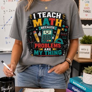 Può includere: Una t-shirt grigio antracite a maniche corte con una stampa grafica colorata che recita I TEACH MATH BECAUSE PROBLEMS ARE MY THING. Il design include una calcolatrice, una matita e un righello in stile cartoon circondati da equazioni.