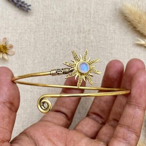 May include: A gold-toned metal cuff bracelet featuring a sunburst design at one end with a central iridescent round stone. The opposite end of the bracelet terminates in a simple spiral coil. The band is thin and flexible with a polished finish.