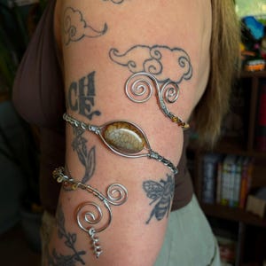 Peut inclure: Un bracelet de bras en fil métallique argenté orné d'une pierre ovale brune à la texture craquelée. Le fil métallique s'enroule autour du bras en spirale, se terminant par deux volutes décoratives enroulées à chaque extrémité de la bande.
