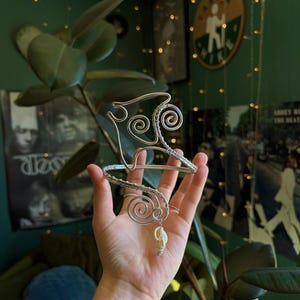 May include: A silver-coloured wire arm cuff featuring intricate spiral designs and a small, off-white seashell charm dangling from the lower section. The metal wire is wrapped tightly to form the structure, creating a continuous, open-ended band.