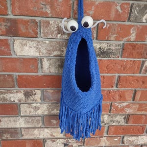 以下が含まれることがあります： 青い毛糸で編まれた、目が飛び出ていてフリンジのついたぶら下がりの玩具。レンガの壁にぶら下がっている。