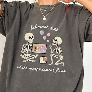 May include: A charcoal grey t-shirt featuring a graphic of two skeletons interacting with a timer and cards. The text reads behaviour goes where reinforcement flows. The skeletons are white with black outlines, and the graphic includes purple bubbles.
