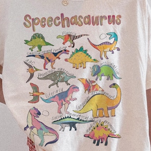 May include: An ivory t-shirt featuring the text "Speechasaurus" and a colorful graphic of various dinosaurs labeled with speech therapy terms like "fluency," "feeding," "articulation," "literacy," "language development," and "social communication."