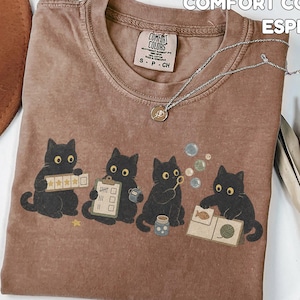 May include: An espresso-coloured short-sleeve t-shirt featuring a graphic of four black cats engaged in various activities, including holding a five-star rating card, a clipboard, blowing bubbles, and looking at a book with a fish and yarn illustration.