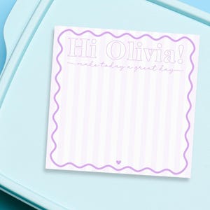 May include: A light blue lunchbox with a matching lid. A white and lavender striped note with the text "Hi Olivia! make today a great day" is on top of the lid. The note has a wavy border and a small heart at the bottom.