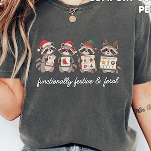 May include: A charcoal gray short-sleeve t-shirt featuring a graphic of four raccoons in holiday attire holding various items. The text reads 'functionally festive & feral' below the raccoons. The top right corner displays 'COMFORT COLORS PEPPER'.