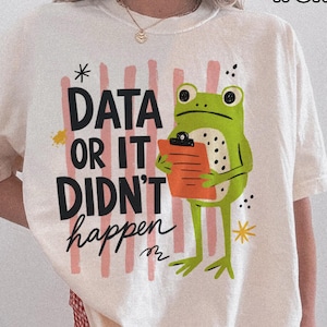 May include: An ivory short-sleeve t-shirt featuring a graphic of a green frog holding an orange clipboard. The text reads "DATA OR IT DIDN'T happen" in black letters over vertical pink brush strokes. The shirt is paired with red and white gingham shorts.