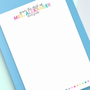 以下が含まれることがあります： 青い縁取りのある白いメモ帳で、カラフルなタイトルが「from the desk of MRS. ALEXANDER third grade」と書いてあります。メモ帳の下の縁は、カラフルな星が並んだ列で飾られています。