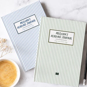 May include: Two rectangular reading journals with vertical stripes. One journal is light blue and white, the other is sage green and white. Both feature a central label with the text "MEGHAN'S READING JOURNAL" and "my 2021 book log".
