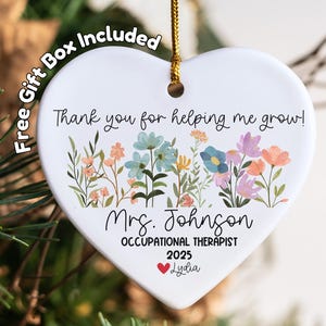 May include: A heart-shaped ceramic ornament with a floral design and the text "Thank you for helping me grow!" It includes the name "Mrs. Johnson", "OCCUPATIONAL THERAPIST", the year "2025", and the name "Lydia". A gold string is attached for hanging.