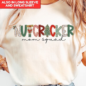 May include: Ivory-colored t-shirt with a festive "NUTCRACKER mom squad" design. The text features a nutcracker, Christmas tree, and various patterns in red, green, and black. The shirt is available in long sleeve and sweatshirt styles.