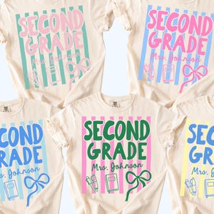May include: Cream-coloured t-shirts with the words "SECOND GRADE" in various colours, including pink, blue, green, and yellow. Each shirt has a teacher's name, a bow, and school supplies. The shirts are labelled "COLOR 1" through "COLOR 5".