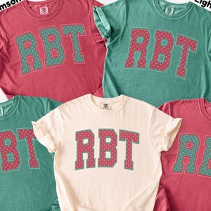 May include: Five short-sleeved crew neck t-shirts in crimson, light green, and ivory colors. Each shirt features the letters RBT in a collegiate font across the chest. The letters are filled with a small polka dot pattern and outlined in a contrasting color.