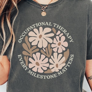 May include: A gray t-shirt with a graphic design of a circle with the text "Occupational Therapy Every Milestone Matters" and stylized flowers in pink and brown.