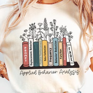 May include: A white t-shirt featuring a graphic of a bookshelf with books representing different aspects of applied behaviour analysis. The books are labelled "PROMPT", "Replacement Behaviour", "Positive Reinforcement", "Data Collection", "Frequency", "Behaviour Reduction", "Function", "Measurement", and "FADE". The text "Applied Behaviour Analysis" is printed below the bookshelf.
