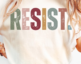 May include: Cream-colored t-shirt with the word "RESIST" printed across the front in a distressed, multi-colored font. The letters are in shades of green, pink, burgundy, and gold. The shirt has short sleeves and a relaxed fit.