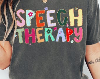 May include: A charcoal gray short-sleeve t-shirt featuring the text SPEECH THERAPY in large, multi-colored, outlined letters. The design includes small decorative icons like flowers, stars, and hearts. The text COMFORT COLORS PEPPER appears in the top right corner.