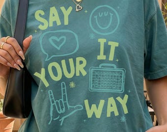 May include: A blue spruce colored t-shirt featuring the text "SAY YOUR IT WAY" in light green. The graphic includes a heart in a speech bubble, a smiley face, a hand gesture, and a small crate. The text "COMFORT COLORS BLUE SPRUCE" is visible.