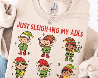 May include: Beige Gildan Sand sweatshirt with the text "JUST SLEIGH-ING MY ADLS" in red. The design features cartoon elves in green outfits, red and green hats, and various accessories. A silver necklace is draped across the sweatshirt.