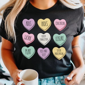 May include: A black t-shirt with a graphic of nine colorful hearts, each with a different word related to occupational therapy. The words are: ADLs, IADLs, Education, Work, Health Management, Leisure, Play, Rest and Sleep, and Social Participation.