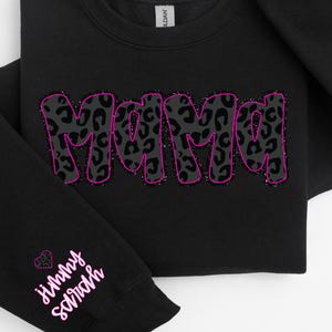 Puede incluir: Sudadera negra con un contorno rosa de la palabra "Mama" en un diseño de estampado de leopardo. La manga tiene un corazón rosa y el texto "jimmy schmidt" en rosa.