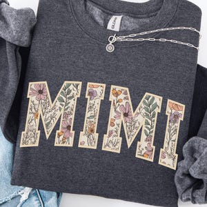 May include: Dark grey sweatshirt featuring the word "MIMI" in a floral design. A silver necklace with a sunburst pendant is on the sweatshirt. The text "GILDAN DARK HEATHER" is at the top.