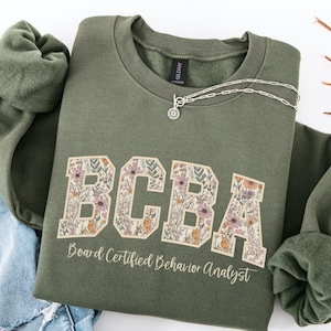 May include: A green sweatshirt with a floral design that spells out "BCBA" and the text "Board Certified Behavior Analyst".
