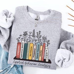 May include: A grey sweatshirt featuring a graphic of a bookshelf with books representing different aspects of Applied Behaviour Analysis. The books are labelled "PROMPT", "Replacement Behaviour", "Positive Reinforcement", "Data Collection", "Frequency", "Behaviour Reduction", "Function", "Measurement", and "FADE".