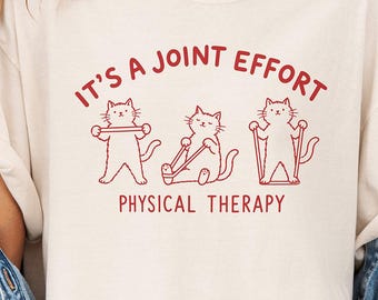 Könnte beinhalten: Ein beiges Kurzarm-T-Shirt mit einer roten Grafik von drei Katzen, die Physiotherapie-Übungen machen. Der Text über den Katzen lautet IT'S A JOINT EFFORT und der Text darunter PHYSICAL THERAPY. Das T-Shirt wird mit einer blauen Jeansjacke getragen.