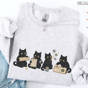 May include: A light gray Gildan Ash crewneck sweatshirt featuring a graphic of four black cats. One cat holds a five-star rating card, another a clipboard, the third blows bubbles, and the fourth examines a book with a fish and yarn illustration.
